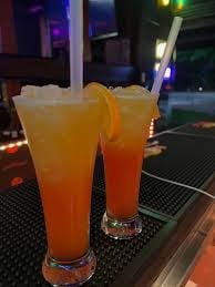 Two amber-colored cocktails with ice in tall glasses on a dark bar counter with colorful lighting in the background