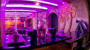Modern nightclub interior with neon purple and pink lighting, featuring bar seating, curved fixtures, and artistic wall decorations